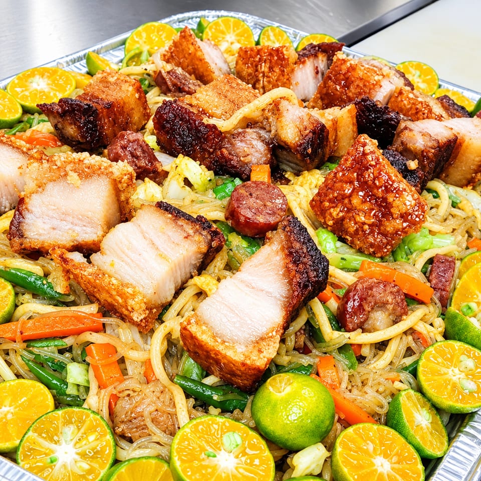 Platter of crispy roast pork, grilled meats, sausage, noodles, vegetables, and sliced calamansi garnishes.