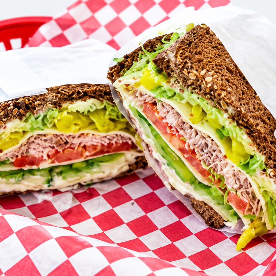 Two halves of a deli sandwich with dark multigrain bread, turkey, cheese, lettuce, tomato, cucumber, and pickles on checkered paper.