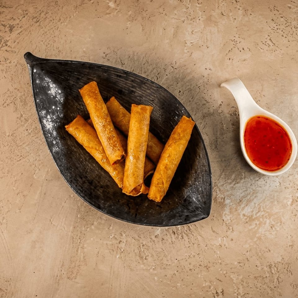 Crispy golden spring rolls served in a black dish with red dipping sauce on a textured stone tabletop.