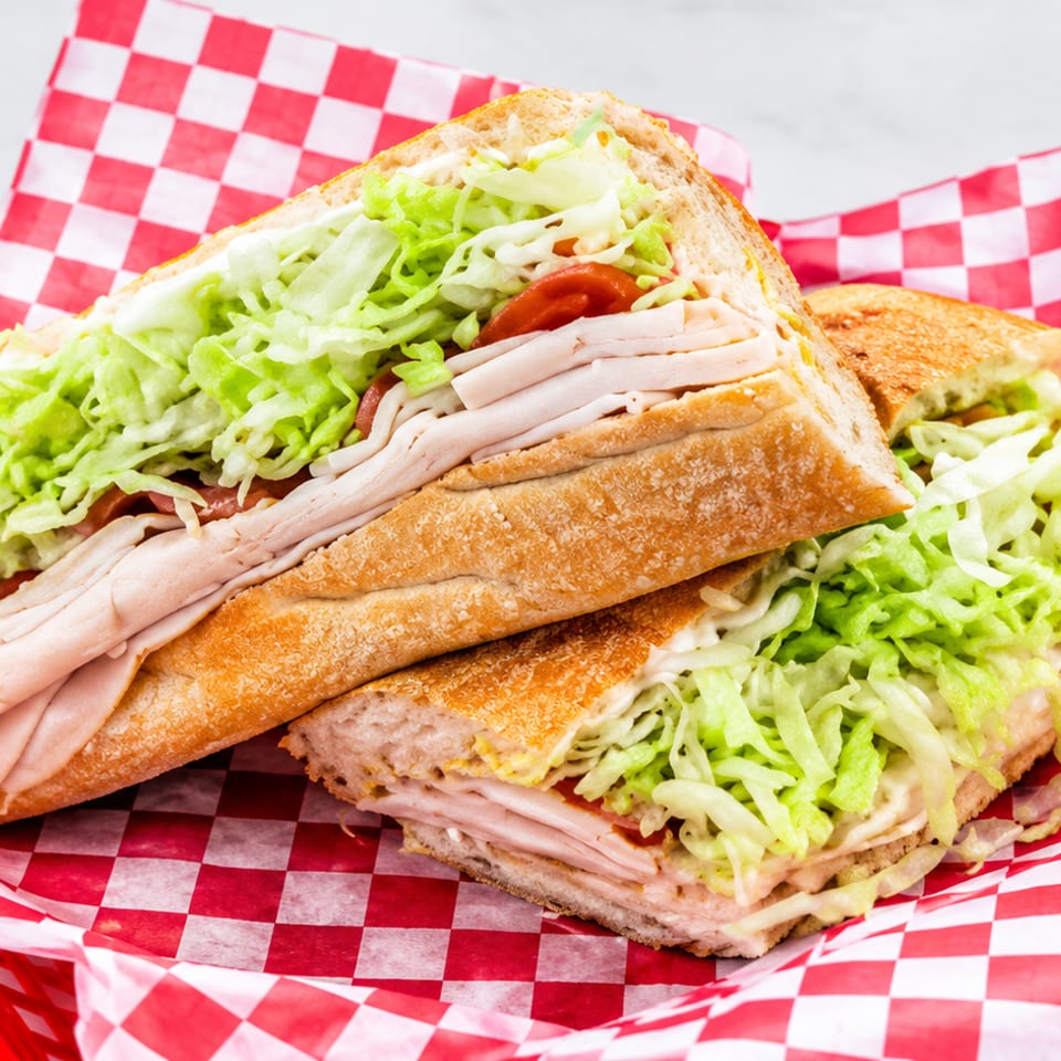 Two turkey and lettuce sandwiches with tomato and mayonnaise on toasted bread, served in a red basket lined with checkered paper.