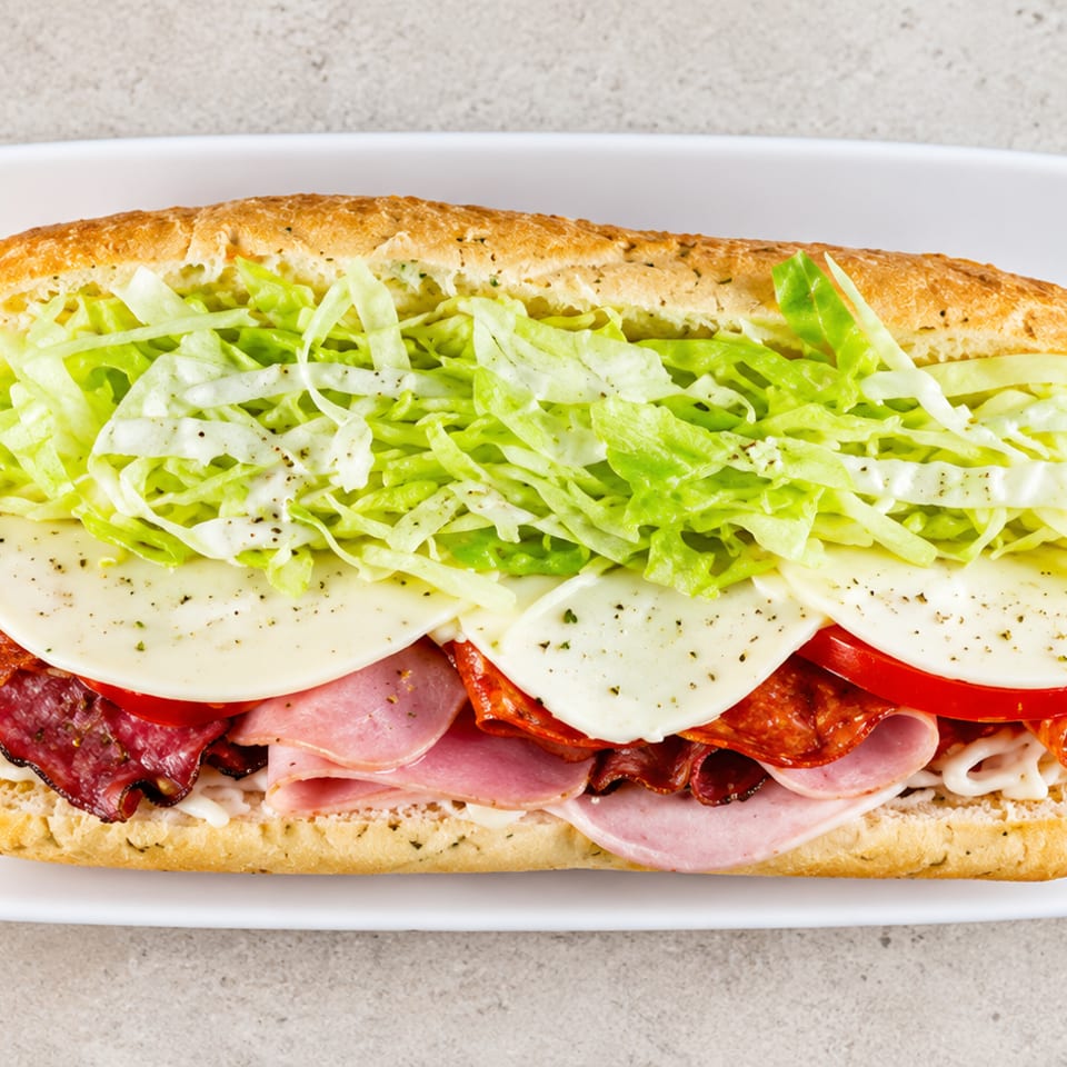 Italian-style sub sandwich layered with ham, salami, pepperoni, provolone, tomato, lettuce, and creamy dressing on a white plate.