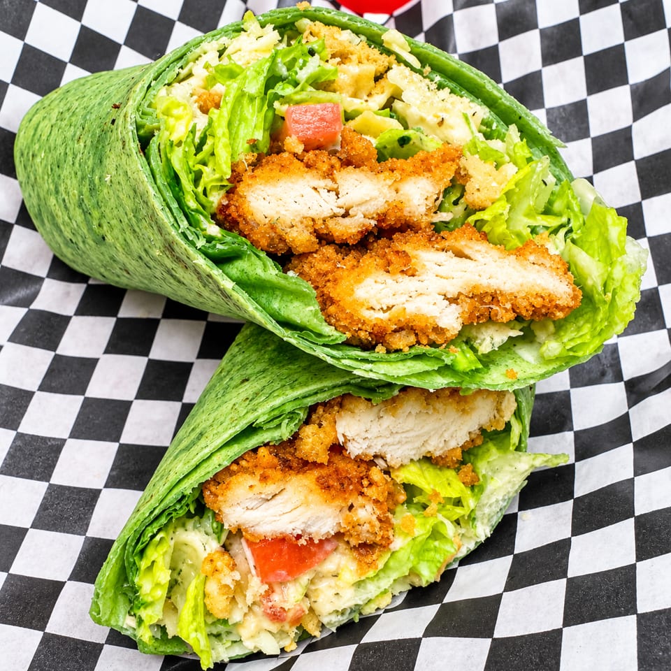 Two spinach wraps filled with crispy chicken, lettuce, tomatoes, and creamy dressing on checkered paper