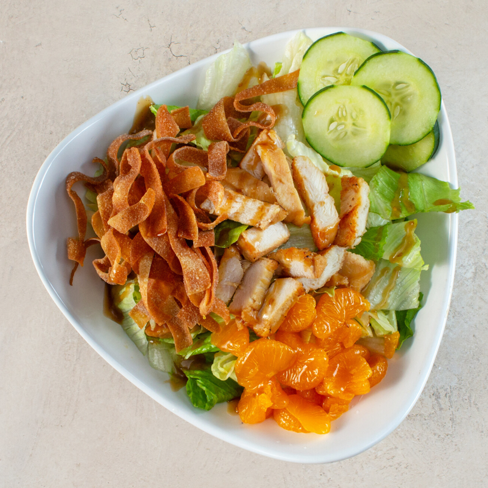 Grilled chicken salad with lettuce, cucumber, mandarin oranges, and crispy tortilla strips in a white bowl.